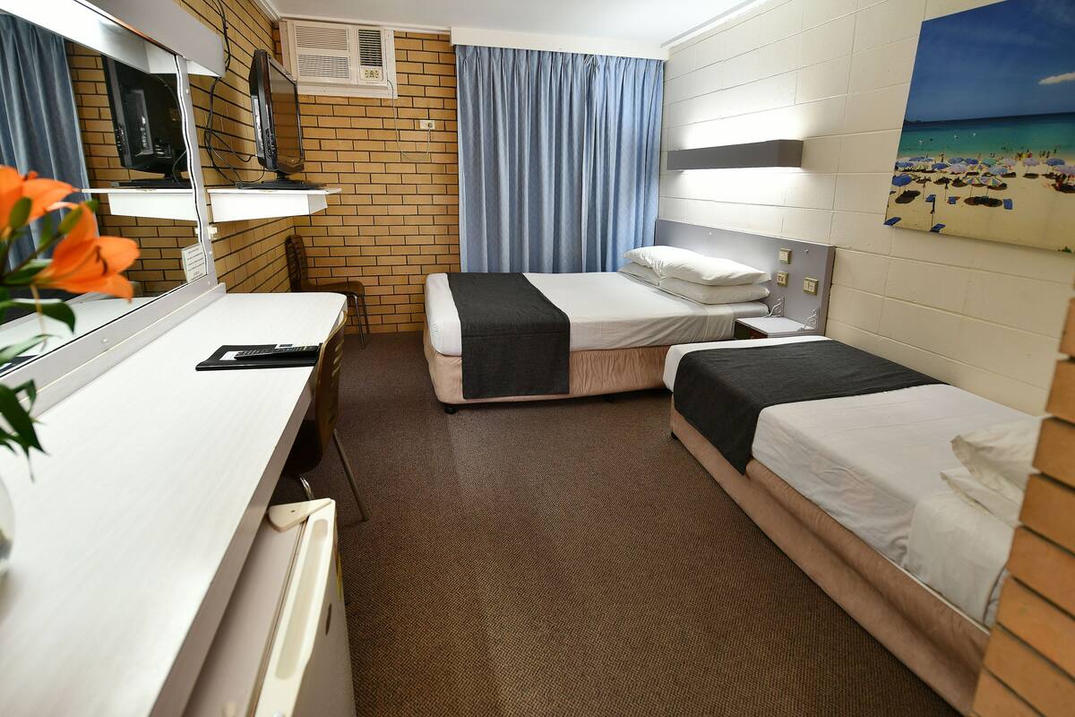 Una habitación de hotel con dos camas individuales, un escritorio con computadora, un espejo y un cuadro con temática de playa en la pared.