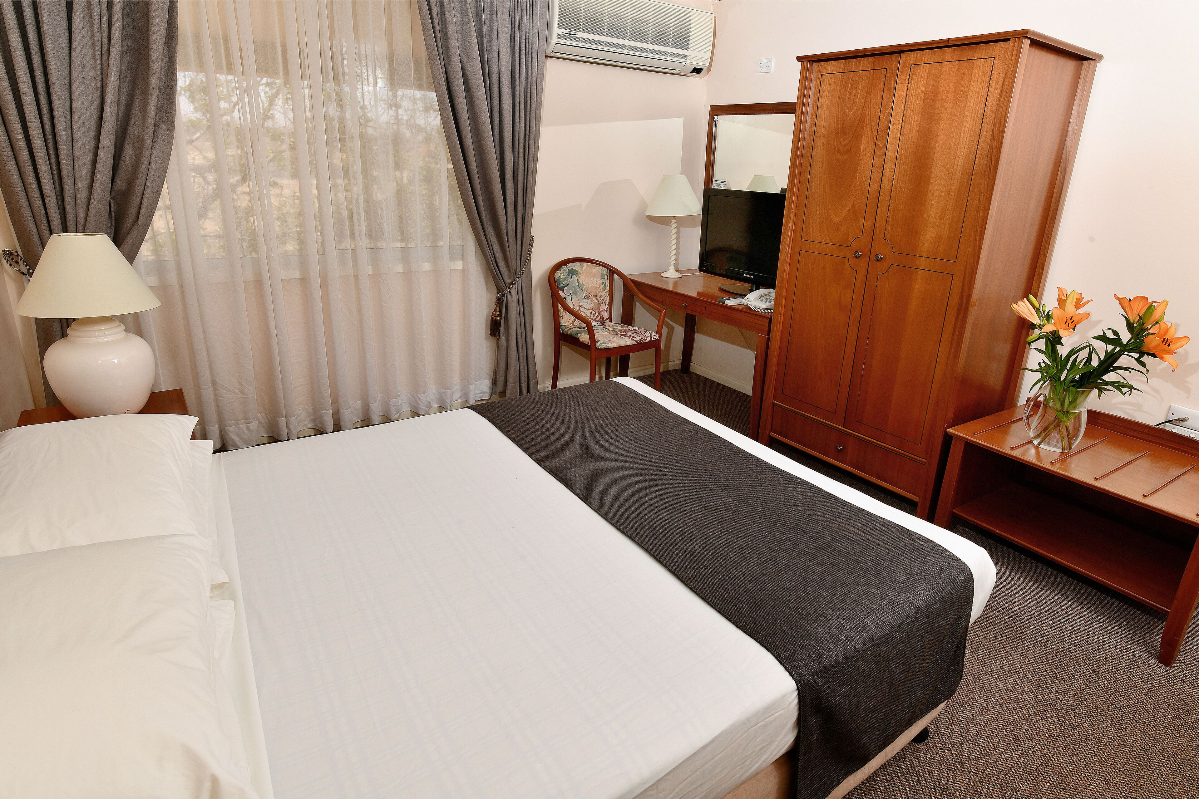 A hotel room with a bed, a desk with a chair, a wardrobe, a lamp, and a vase with flowers.