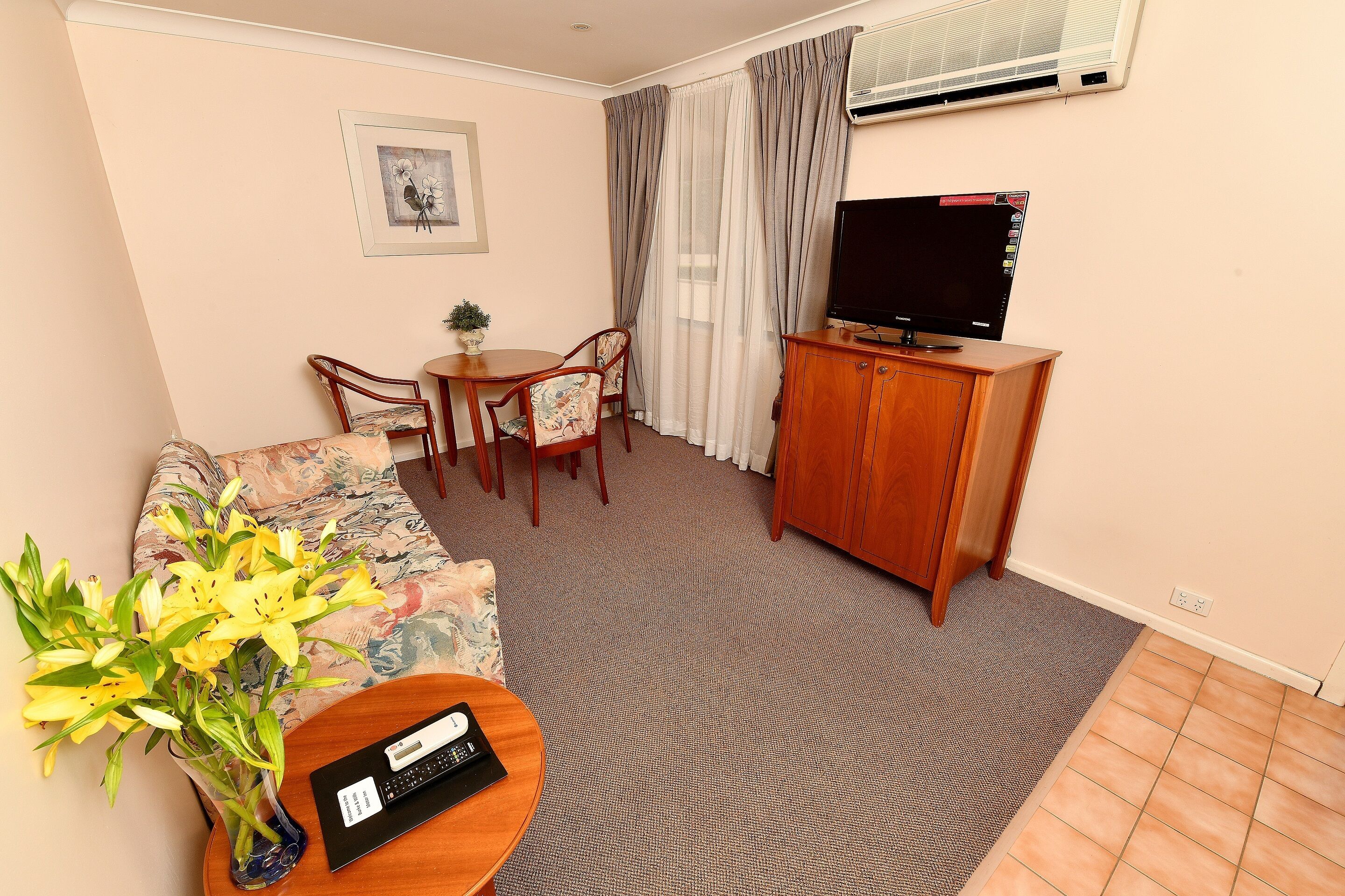 A living room with a sofa, a dining table with chairs, a TV on a wooden cabinet, and a vase of yellow flowers.