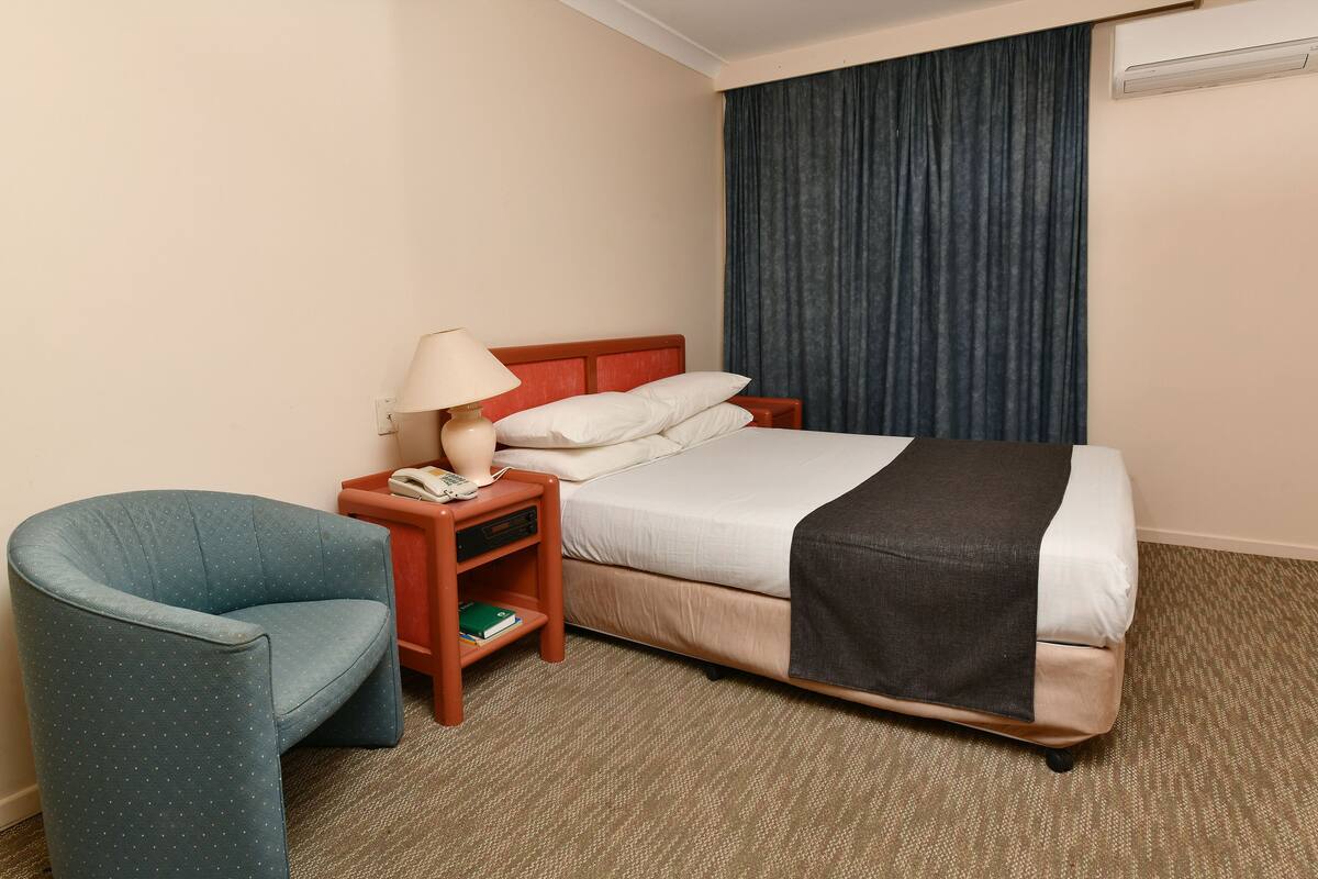 Una habitación de hotel con una cama, una silla, una mesita de noche, una lámpara y una cortina.