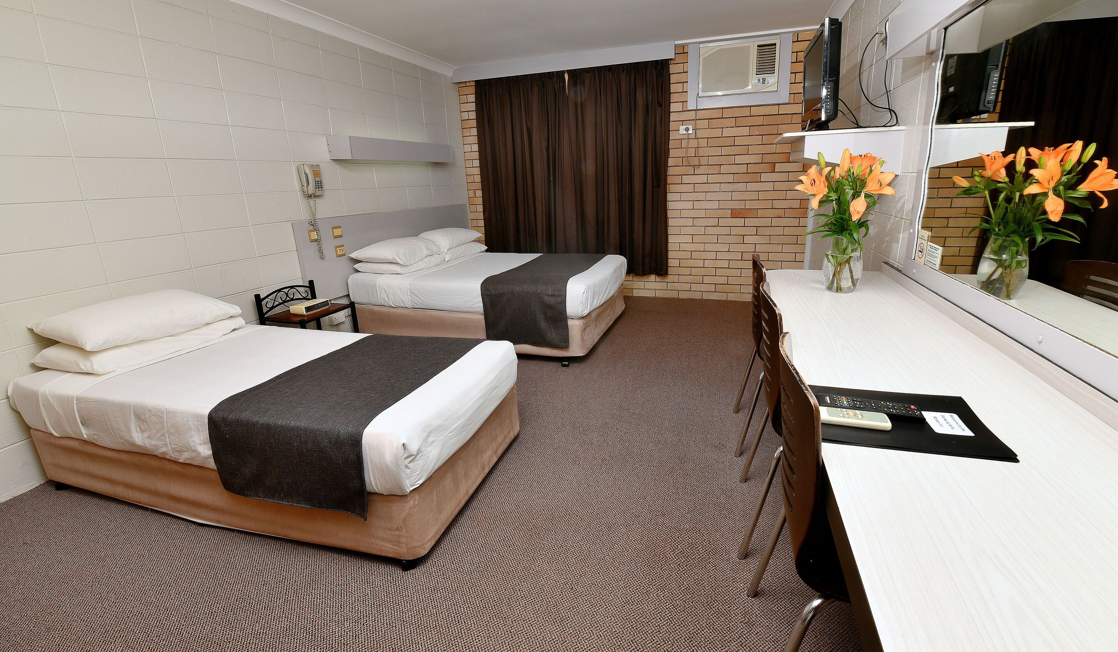 A hotel room with two beds, a desk, a chair, a telephone, a TV, and a vase with flowers.