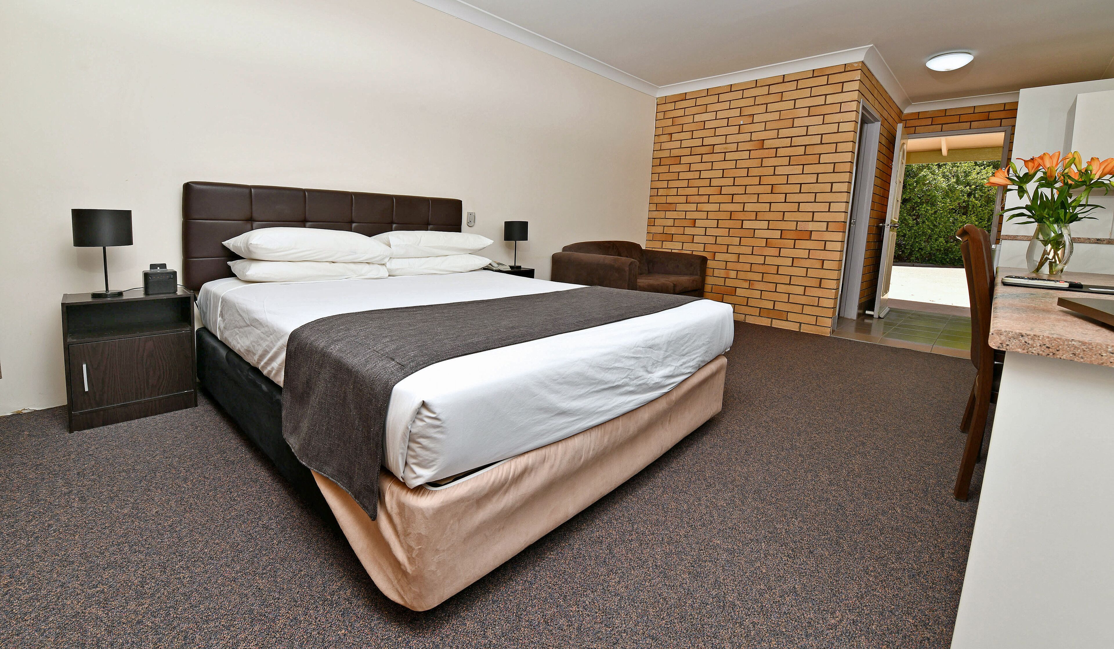 A hotel room with a large bed, a nightstand, a chair, a small table, and a brick wall.