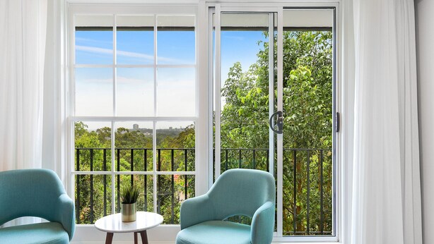A room with a large sliding glass door leading to a balcony with a view of greenery and a cityscape. There are two chairs and a small table with a potted plant. - Killara Hotel & Suites (Killara)