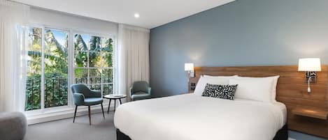 A hotel room with a large bed, two chairs, a small table, and a view of greenery through large windows.