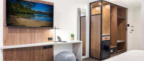 A modern bedroom with a TV mounted on the wall, a desk with a phone, a small plant, and a wardrobe with a mirror.