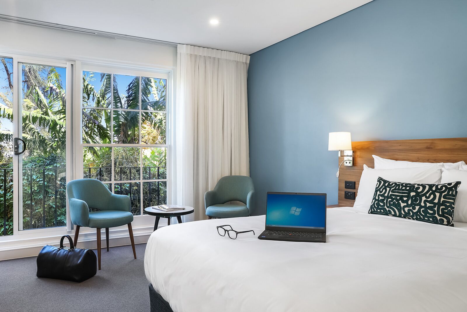 Habitación de hotel con cama, dos sillones, una laptop, una mesita de noche y vistas a follaje tropical a través de amplios ventanales.