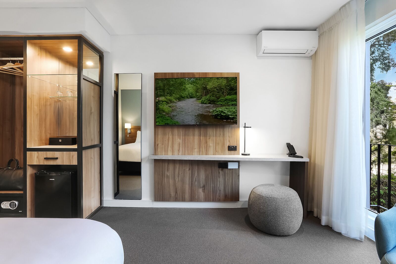 Ett modernt hotellrum med en garderob i trä, en stor spegel, en säng, ett skrivbord med en dator, en ottoman i sten och utsikt över grönområden genom fönstret.