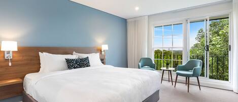 A hotel room with a large bed, two bedside lamps, a view of greenery through large windows, and a balcony with two chairs.