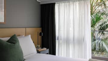 Habitación de hotel con cama, mesita de noche, lámpara y vistas a zonas verdes a través de cortinas translúcidas.