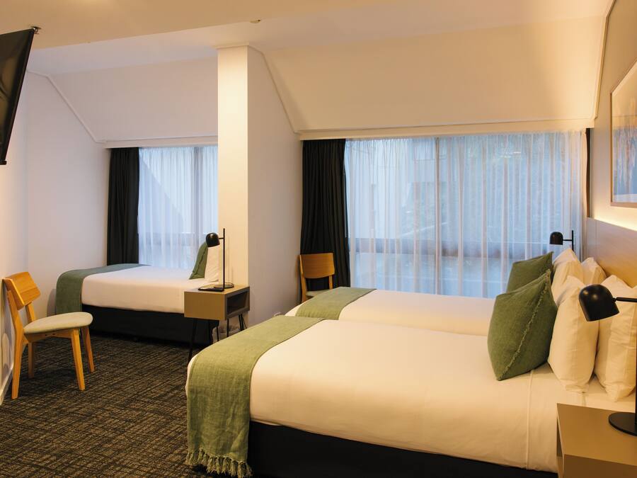 Een hotelkamer met twee bedden, een bureau, een stoel en een groot raam met dunne gordijnen.