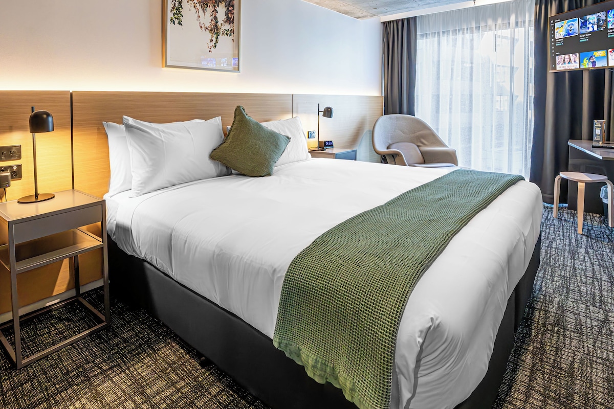 Una habitación de hotel con cama, escritorio, silla, televisor y ventana con cortinas.