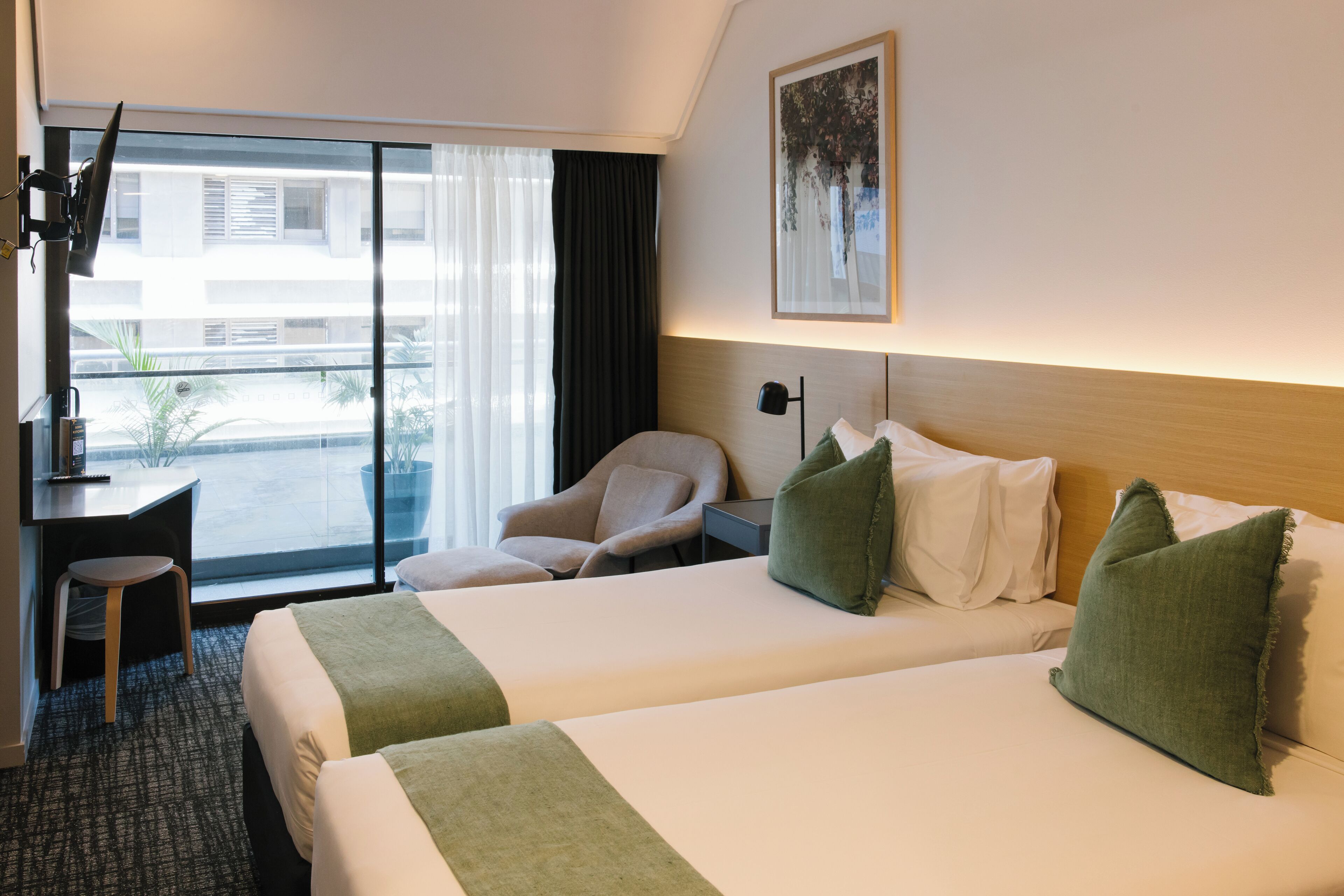 Hotellihuone, jossa on kaksi sänkyä, työpöytä, tuoli ja suuri ikkuna, josta on näkymä ulos.