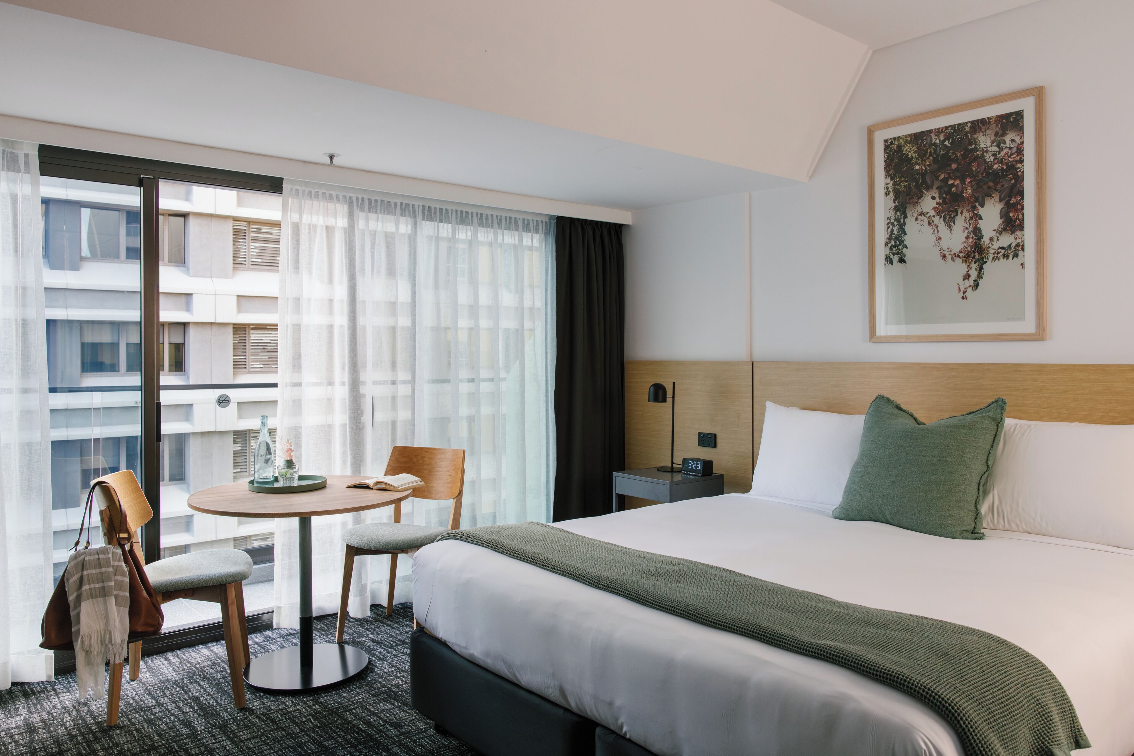 A hotel room with a large bed, a round table with chairs, a small desk, and a window with sheer curtains.