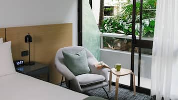 A hotel room with a bed, a grey armchair with a green pillow, a small table with a green mug, a digital clock, and a window with a view of greenery.