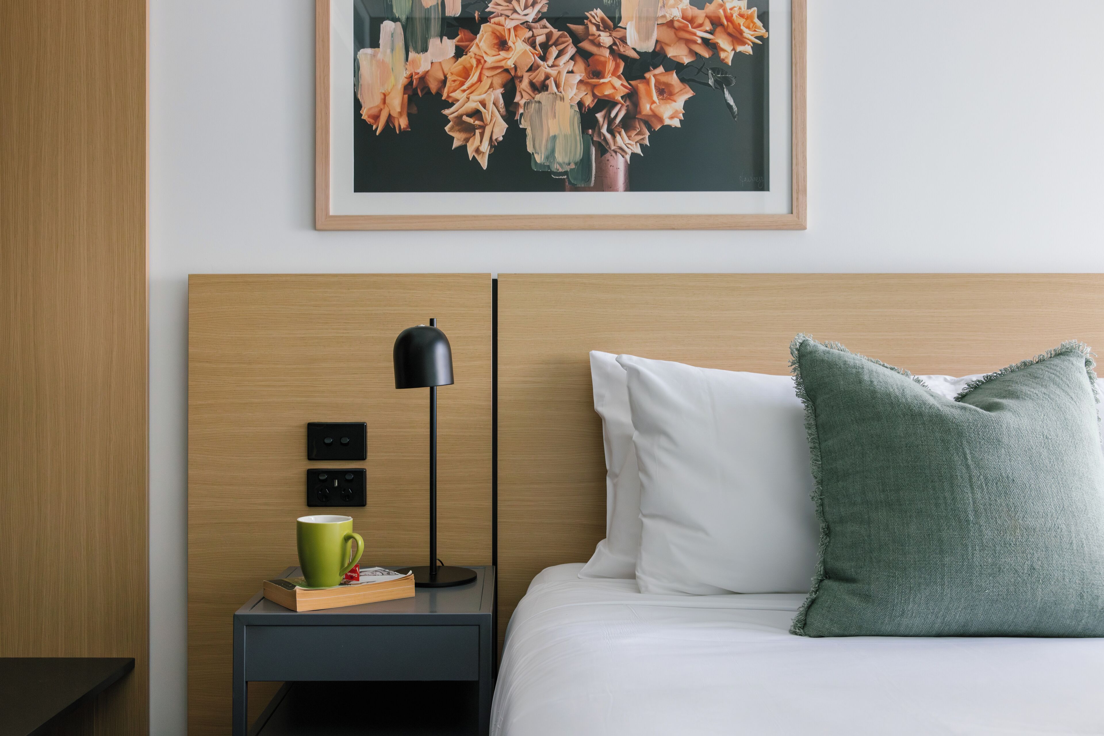 Una habitación de hotel ordenada con cama, mesita de noche con lámpara, taza verde y cuadro con motivos florales en la pared.