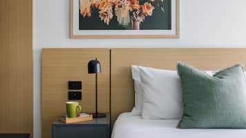Una habitación de hotel ordenada con cama, mesita de noche con lámpara, taza verde y cuadro con motivos florales en la pared.
