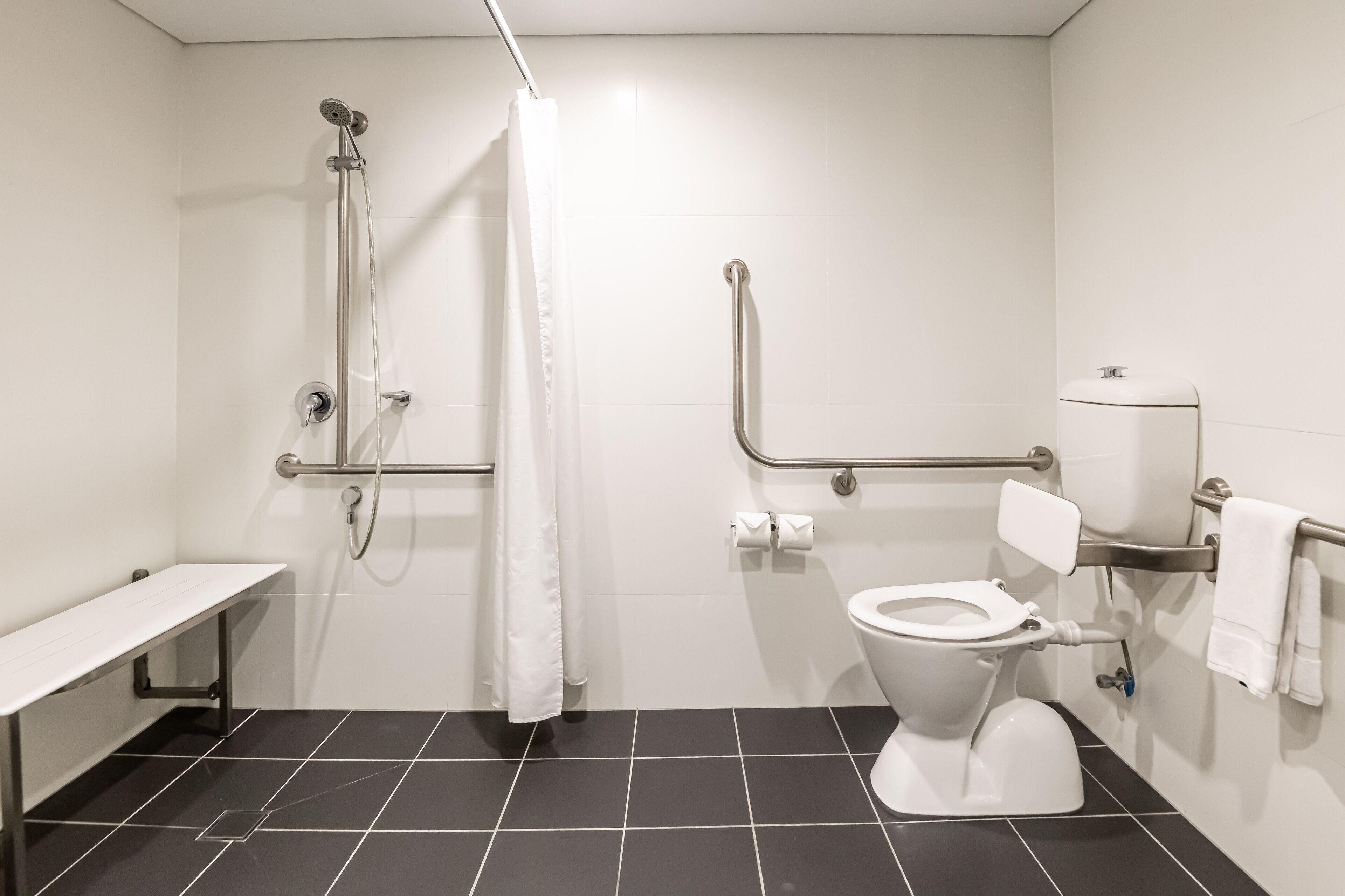 A bathroom with a white toilet, a shower with a grab bar, a bench, and a towel rack.