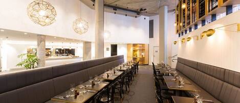Un restaurant doté de longues tables communes et de banquettes, d’un éclairage moderne suspendu et d’une étagère à vin fixée au mur.
