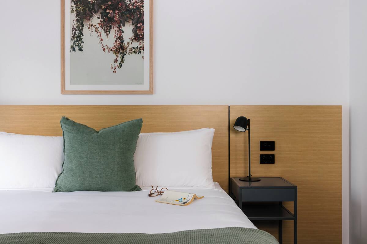 Una cama bien hecha con almohadas blancas y verdes, una obra de arte floral enmarcada encima y una mesita de noche con una lámpara y un libro.