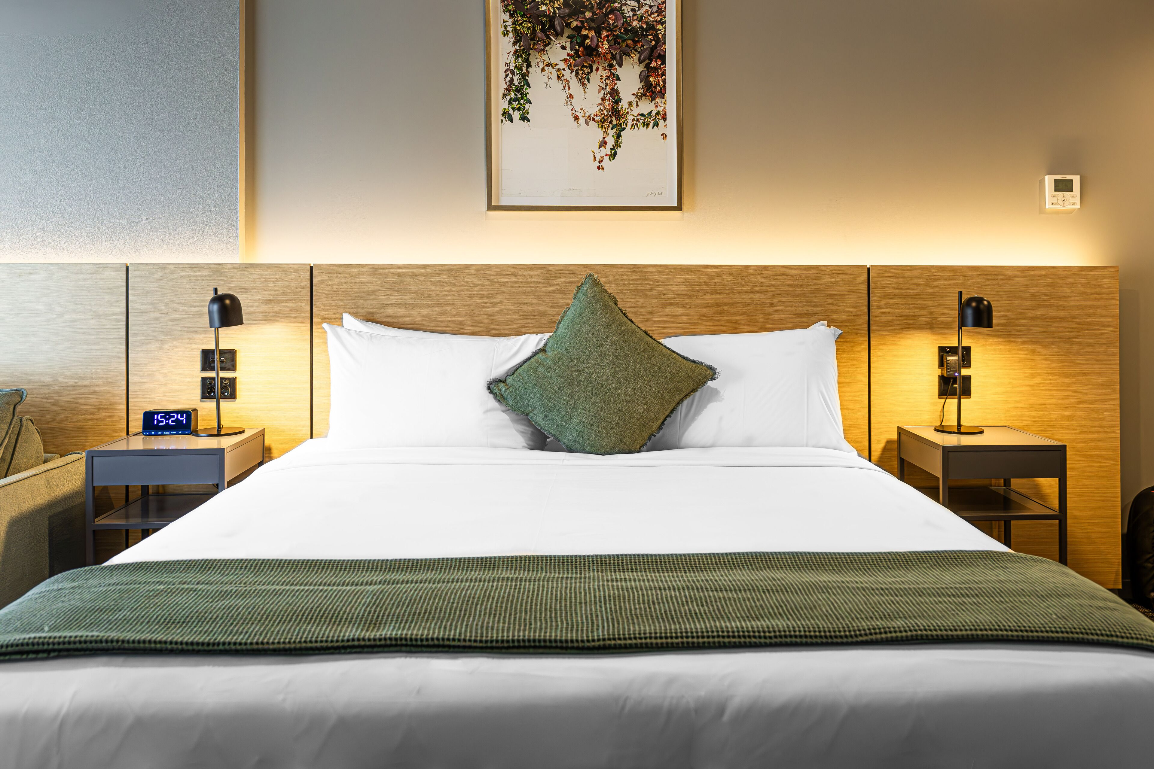 Un lit bien fait, avec un oreiller vert, deux tables de chevet avec des lampes, une horloge et une œuvre d’art accrochée au mur.