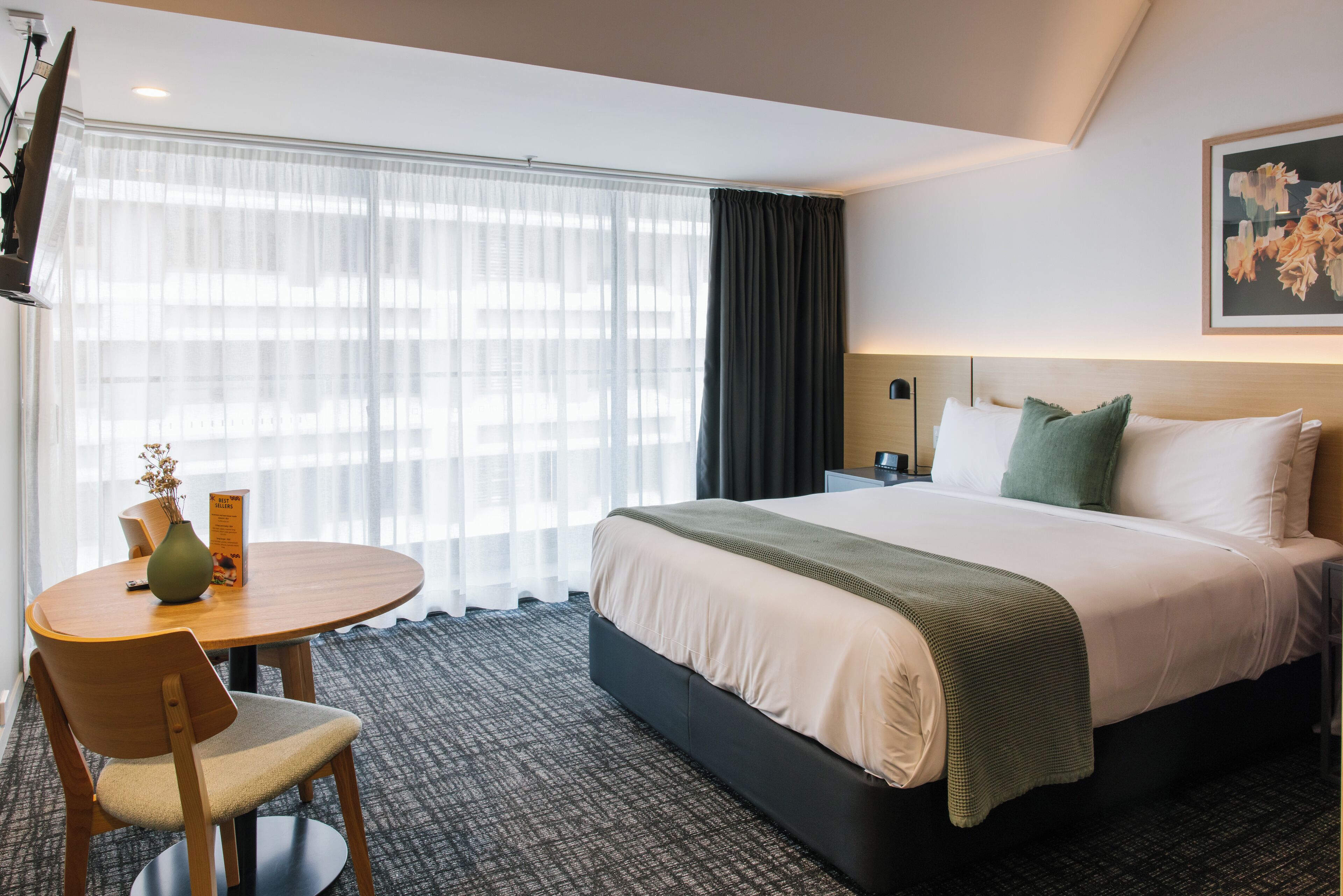 A hotel room with a bed, a round table with a chair, a vase with flowers, and a window with curtains.