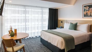 A hotel room with a bed, a round table with a chair, a vase with flowers, and a window with curtains.