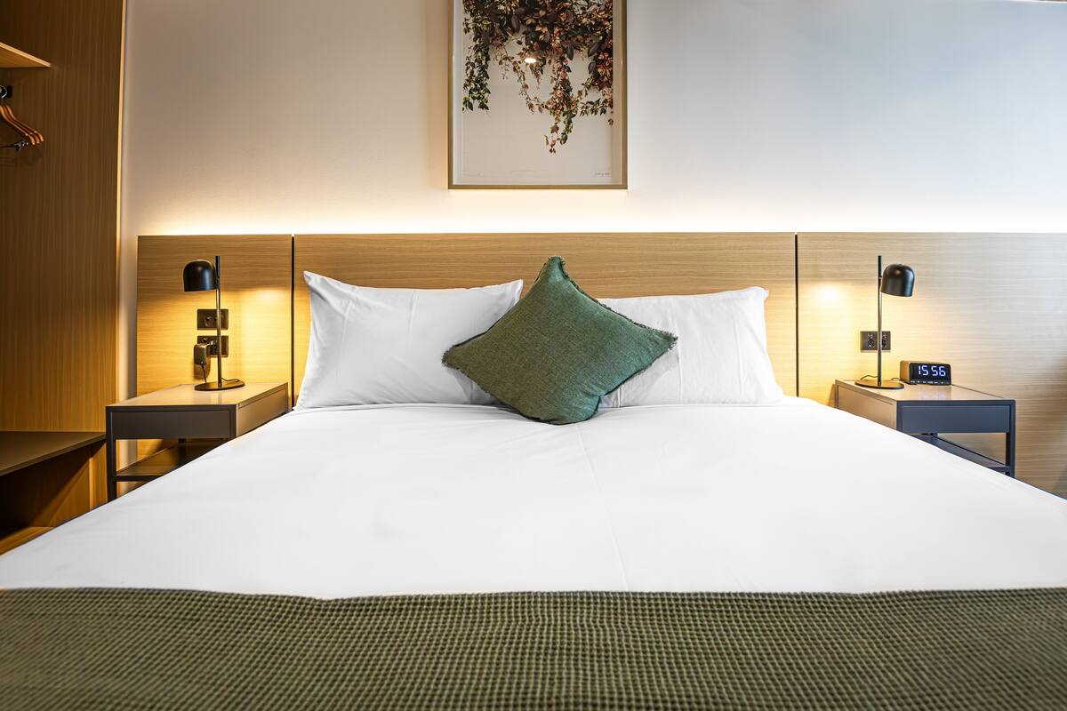 Una cama bien tendida con sábanas blancas y un cojín verde decorativo, flanqueada por dos mesitas de noche con lámparas y relojes.