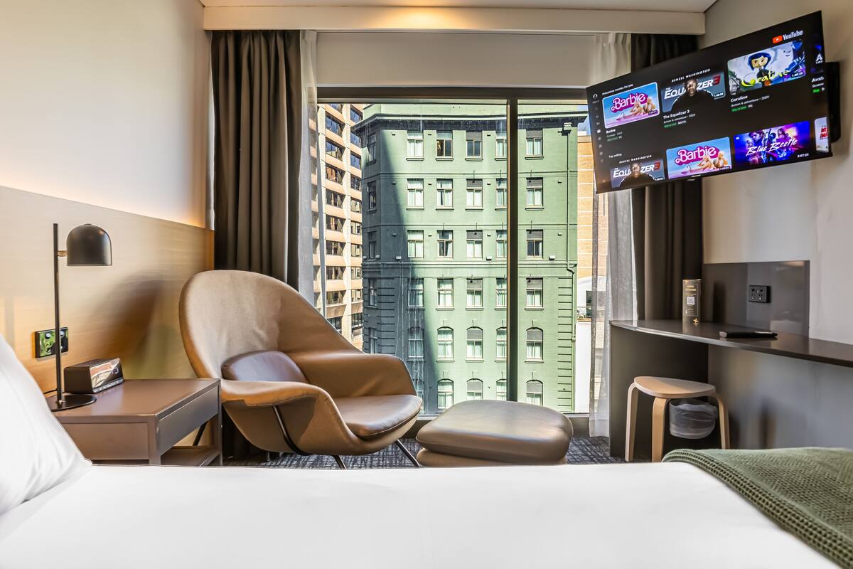 Habitación de hotel con cama, una silla, televisor montado en la pared y vista a un edificio a través del ventanal.