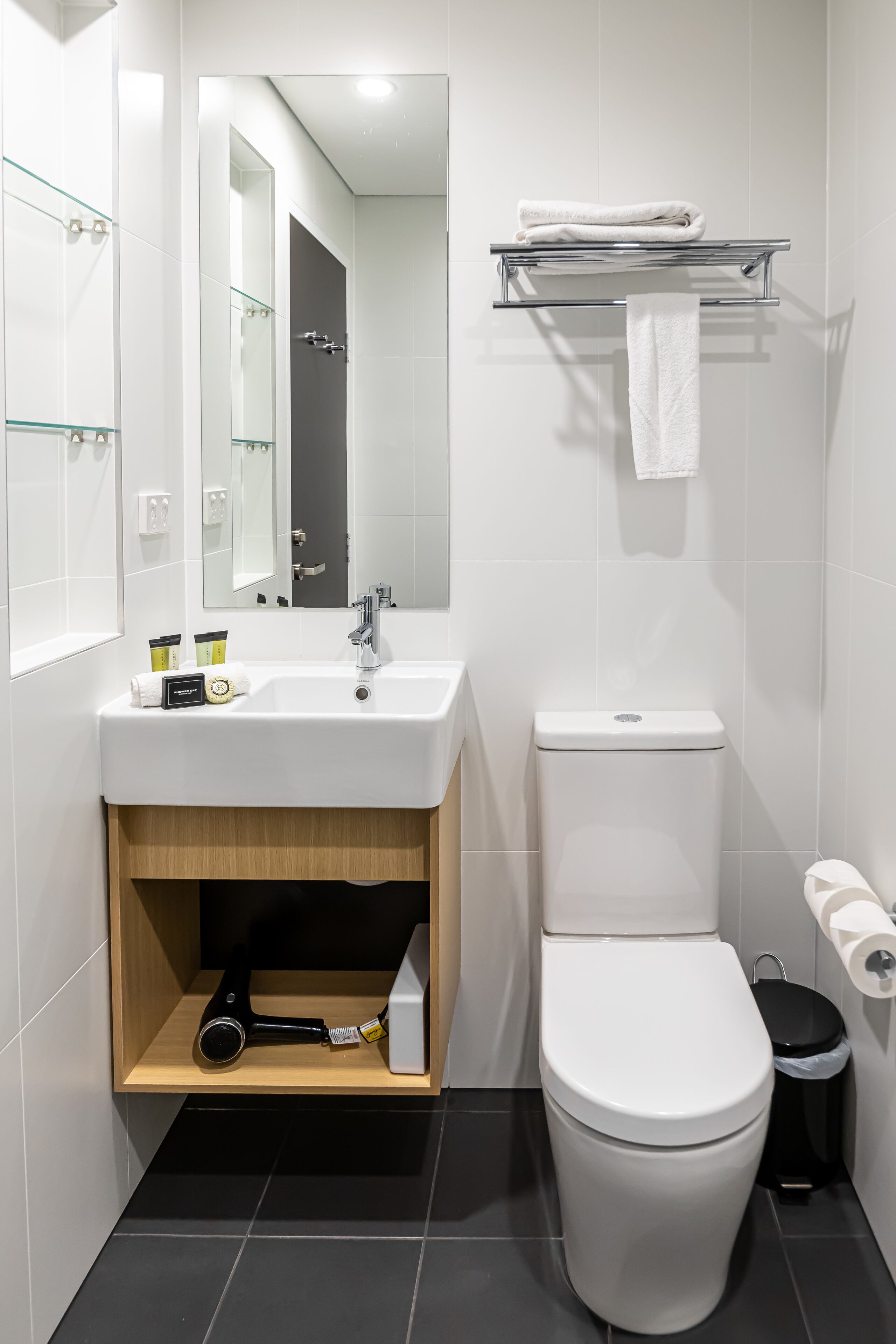Een moderne badkamer met een wit wastafelmeubel, spiegel en toilet. Boven het toilet bevindt zich een handdoekhouder en onder het wastafelmeubel een plank met toiletartikelen.