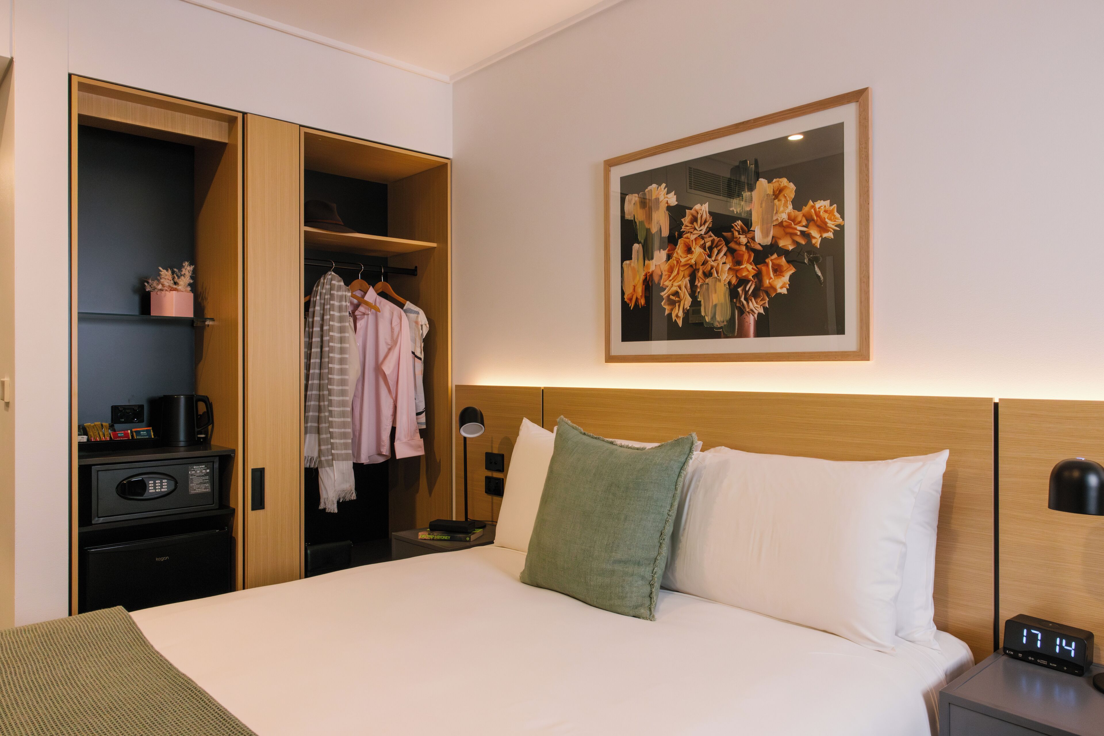 A hotel room with a bed, a nightstand, a desk, a wardrobe, and a framed floral artwork on the wall.