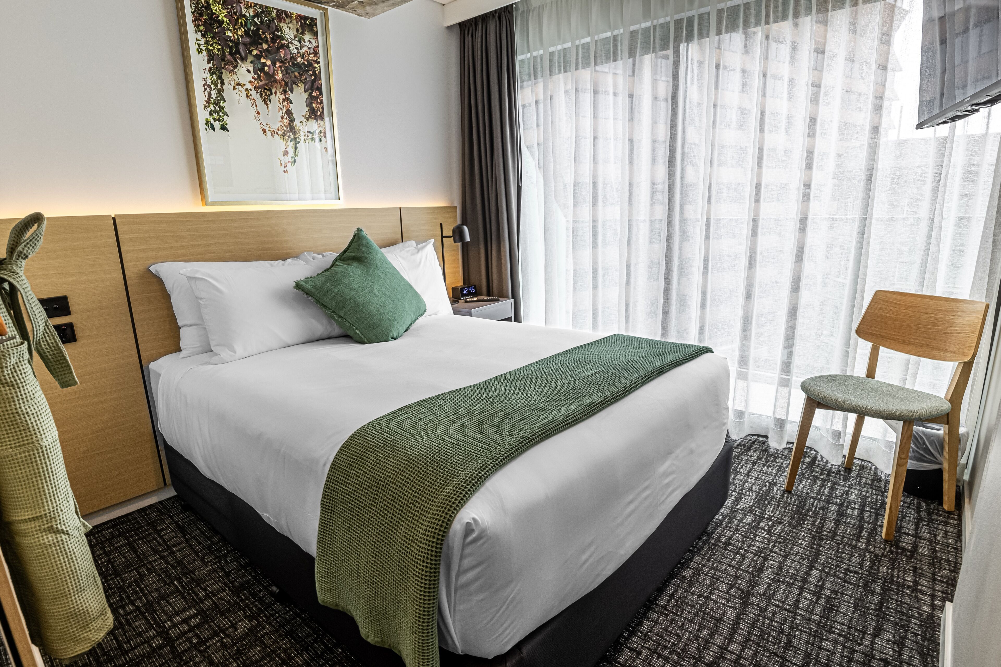 A hotel room with a large bed, a green throw blanket, a wooden nightstand, a chair, and a window with sheer curtains.