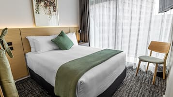 Quarto de hotel com cama grande, manta verde, mesa de cabeceira de madeira, uma cadeira e janela com cortinas leves.
