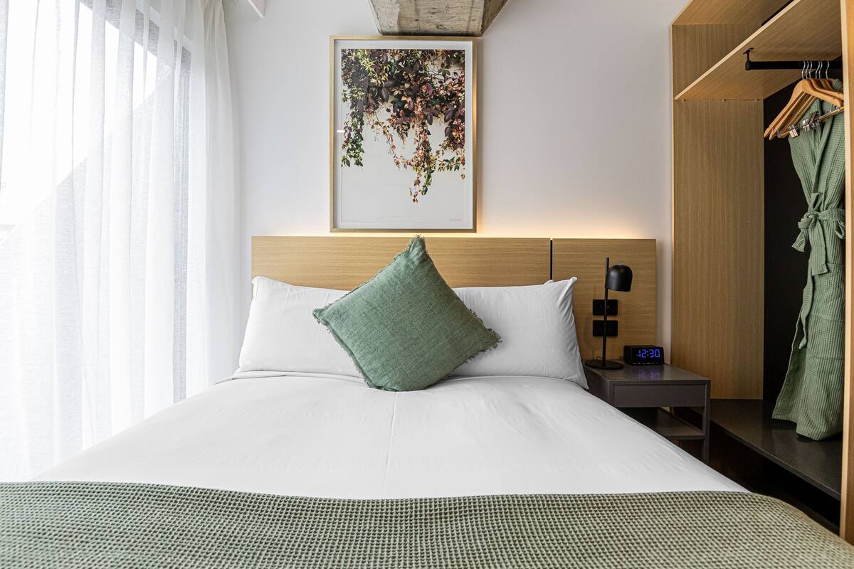 Una cama bien hecha con un cojín verde, una mesita de noche con un reloj y una bata colgada en un perchero.