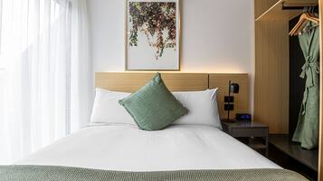 Una cama bien hecha con un cojín verde, una mesita de noche con un reloj y una bata colgada en un perchero.