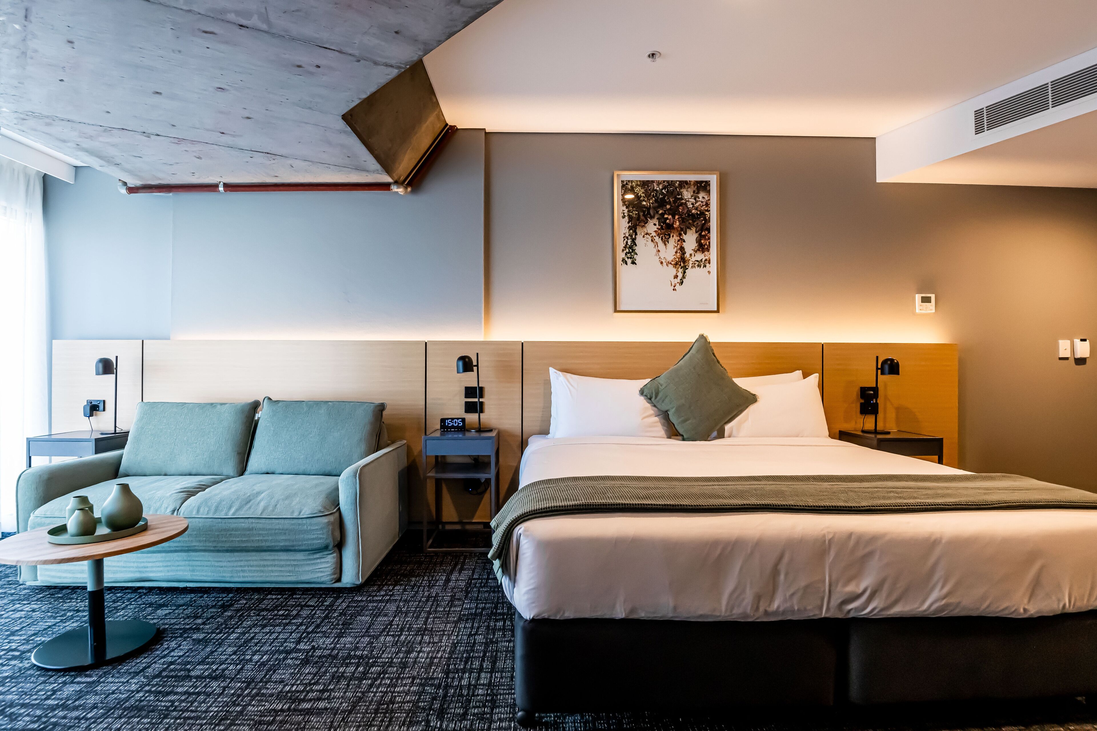 Quarto de hotel com cama, sofá, uma pequena mesa com um vaso, relógio de parede e um quadro emoldurado.