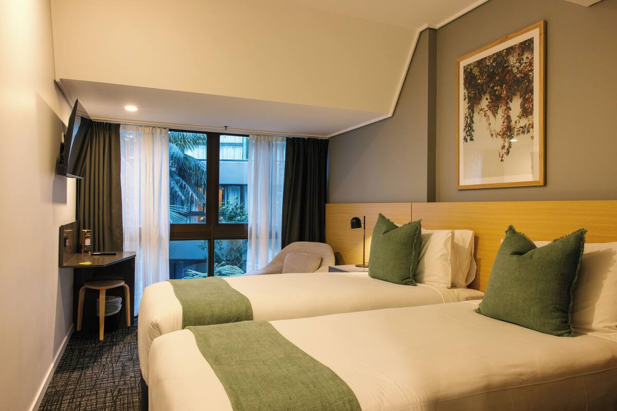 Habitación de hotel con dos camas, un escritorio, una silla, una ventana con cortinas y un cuadro en la pared.