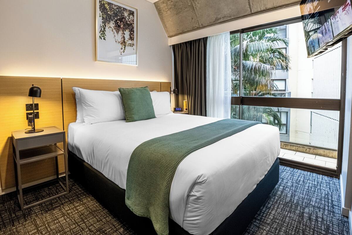 Habitación de hotel con una cama, una mesita de noche con lámpara, una manta verde y un cuadro en la pared, además de vistas a palmeras a través de la ventana.