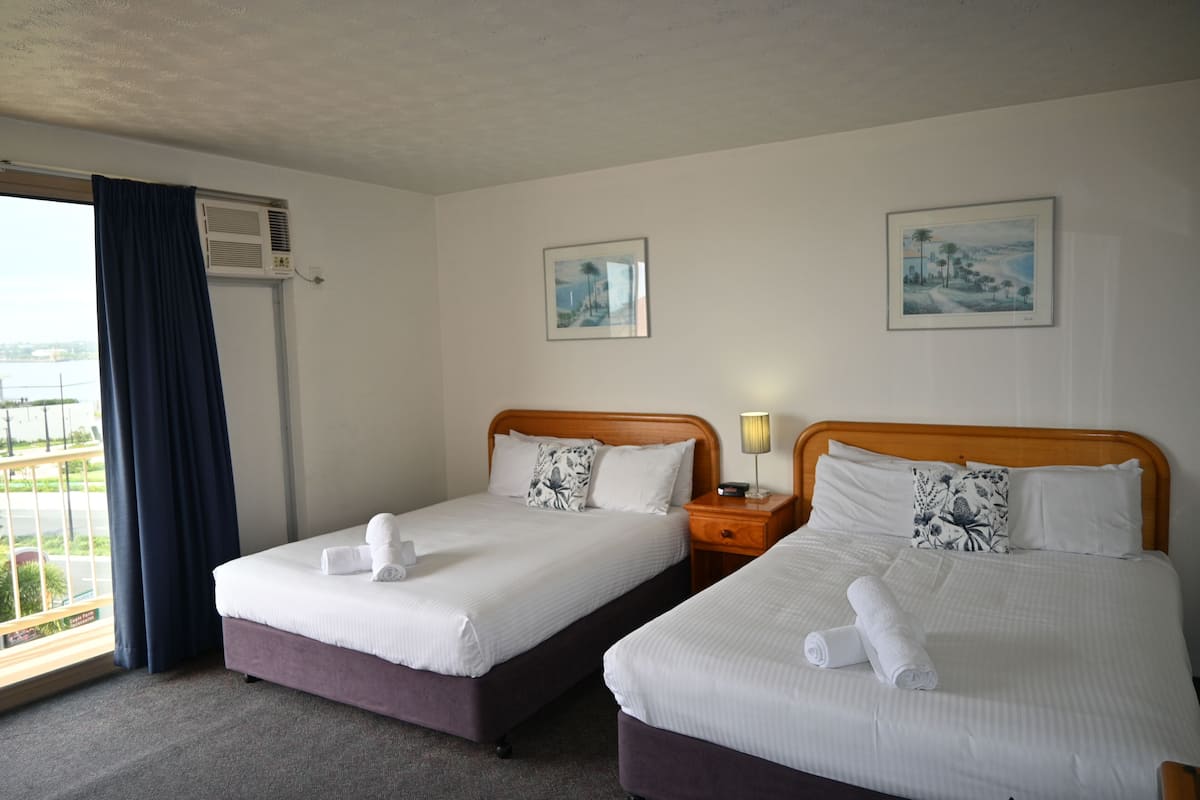 Habitación de hotel con dos camas, una ventana con cortinas, un equipo de aire acondicionado y dos cuadros en la pared.