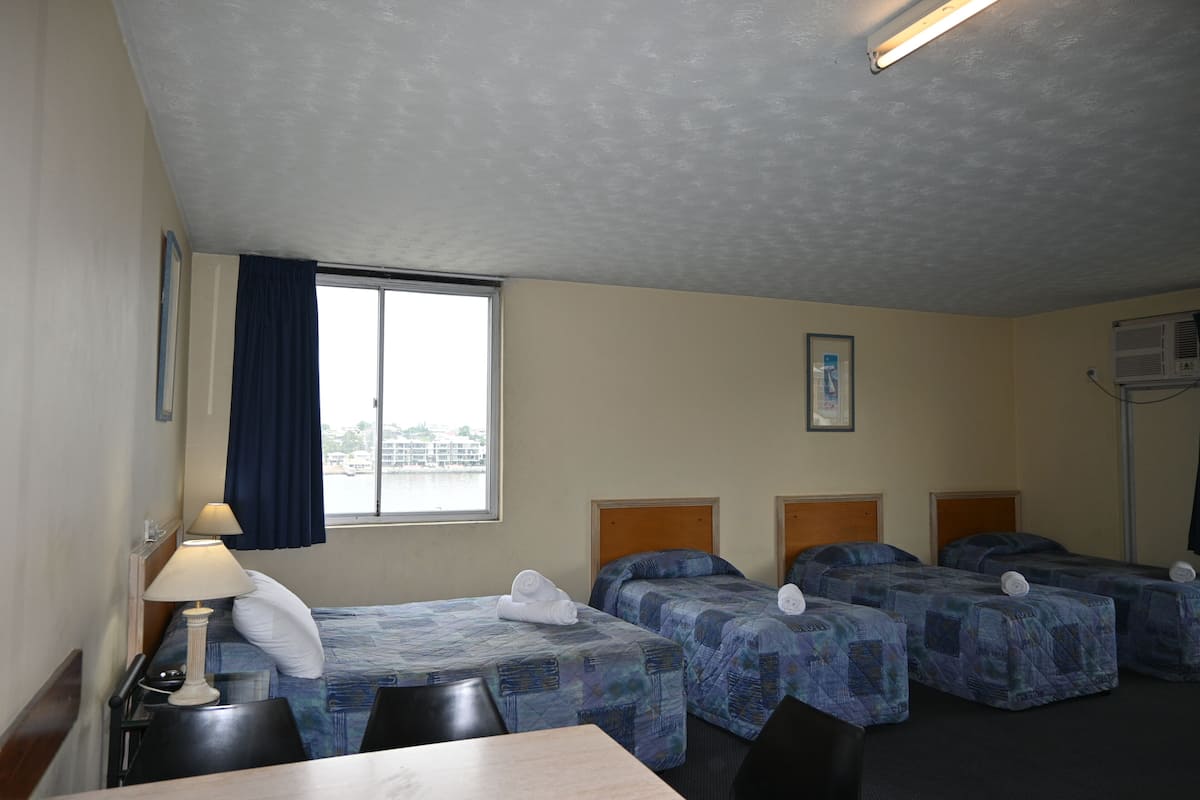 Habitación de hotel con cuatro camas, un escritorio, una silla, una lámpara y ventana con vista al agua.
