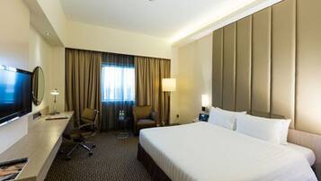 A hotel room with a large bed, a desk with a flat-screen TV, a chair, a small table, and a window with curtains.