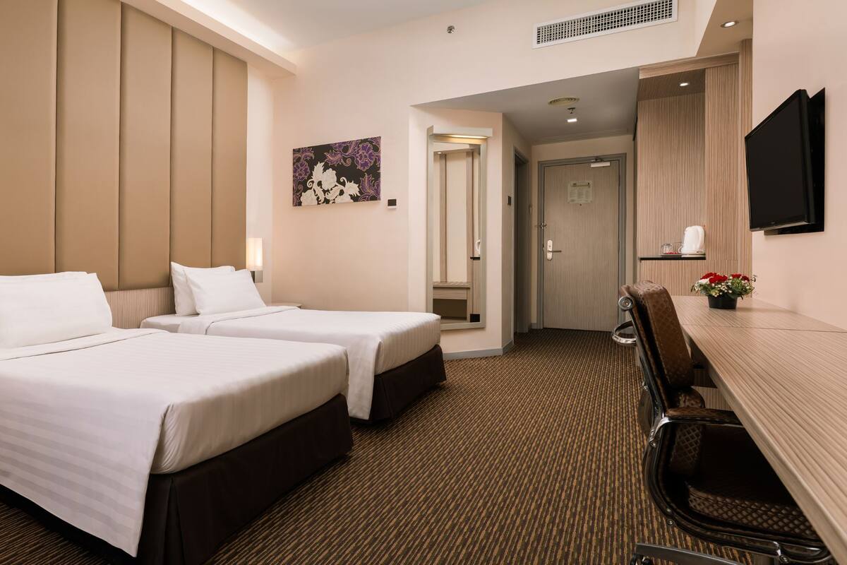 A hotel room with two beds, a desk with a chair, a flat-screen TV, and a small table with flowers.