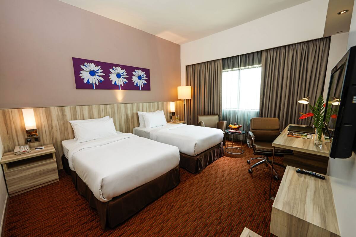 A hotel room with two beds, a desk with a chair, a TV, a lamp, and a painting on the wall.