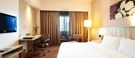 A hotel room with a bed, desk, chairs, a TV, and a window with curtains.
