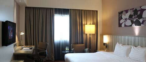 A hotel room with a bed, desk, chairs, a TV, and a window with curtains.