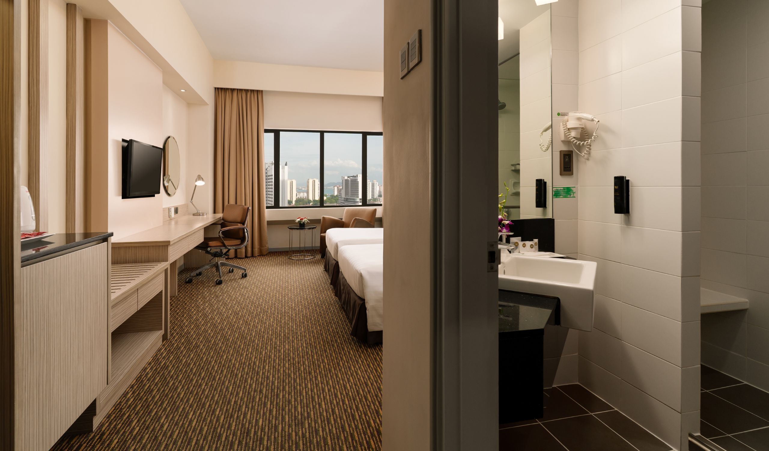 Habitación de hotel con escritorio, dos camas, baño con lavamanos y espejo, y ventana grande con vista a la ciudad.