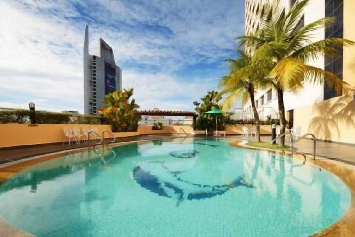 Uma piscina com centro azul cristalino, rodeada por palmeiras e um edifício com design arquitetônico singular ao fundo.