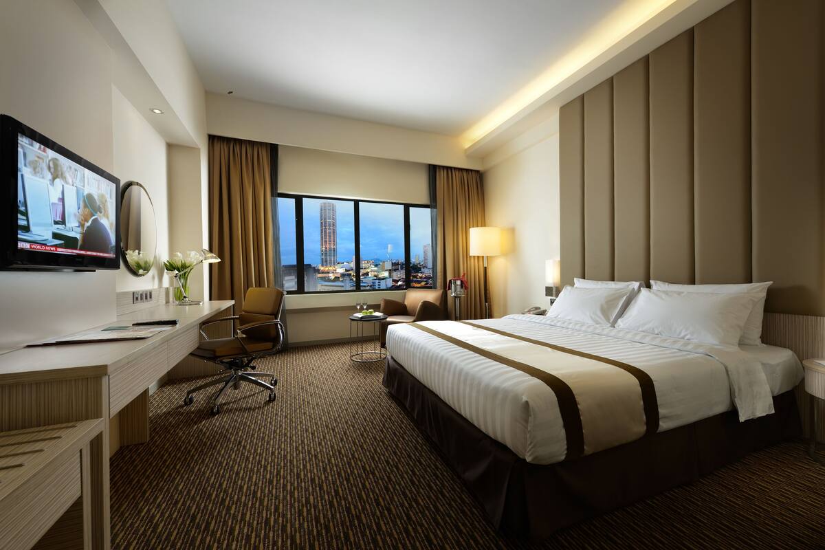 Quarto de hotel com uma cama grande, uma escrivaninha com computador, uma cadeira, um abajur e vista para a cidade pela janela.
