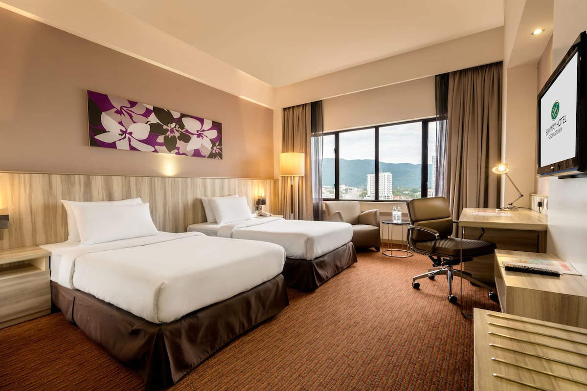 Quarto de hotel com duas camas, uma escrivaninha, uma cadeira, uma televisão e uma janela com vista para a cidade.