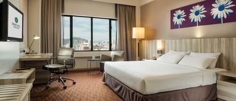 A hotel room with a large bed, a desk, a chair, a small table, and a window with a city view.