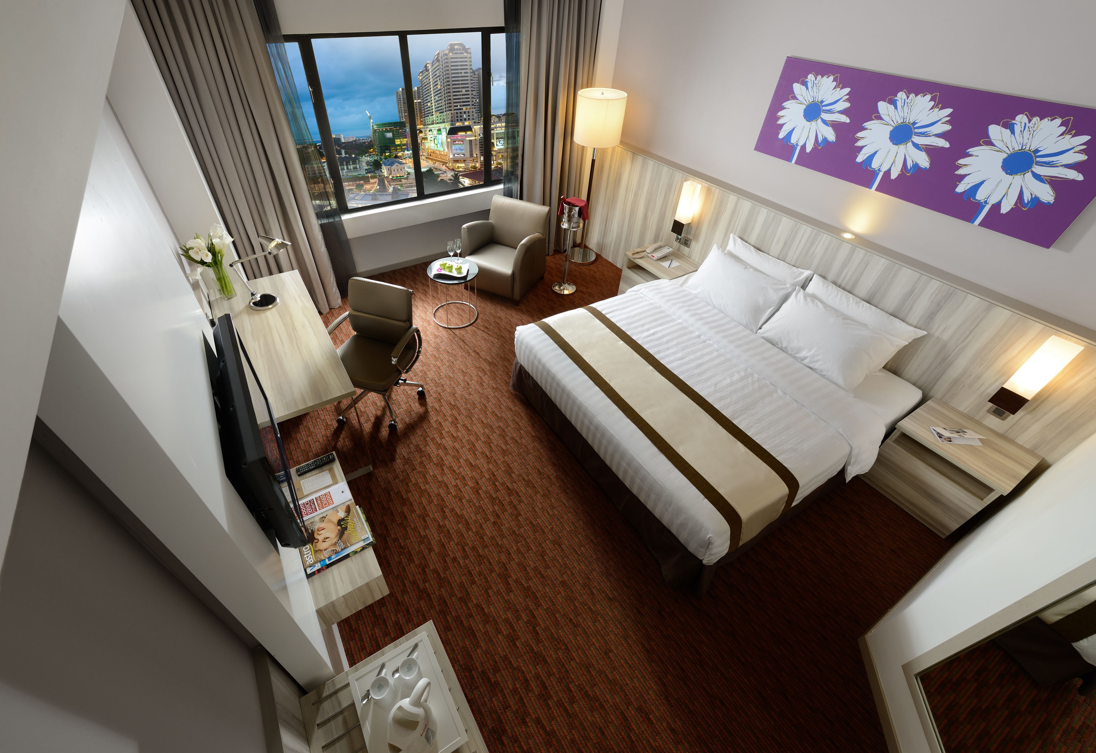 Habitación de hotel con una cama grande, un escritorio con lámpara, una silla y vistas a un paisaje urbano a través de un ventanal amplio.